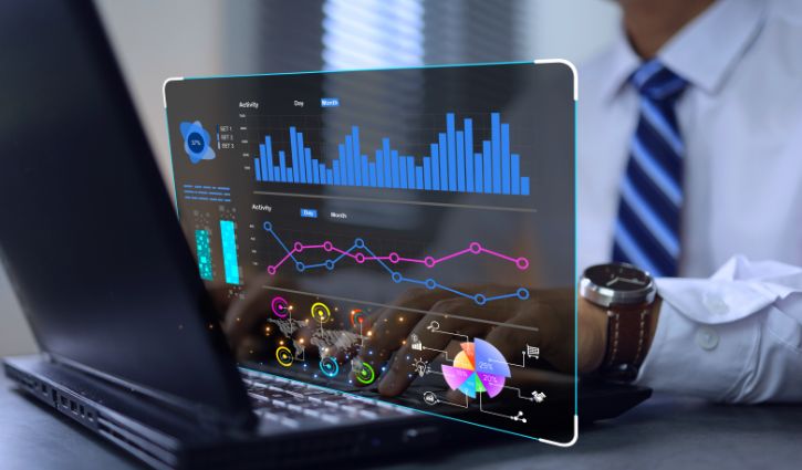 A person in a shirt and tie works on a laptop featuring a futuristic, transparent screen. The screen showcases a management dashboard with vibrant data visualizations, including bar charts, line graphs, and pie charts—ideal for making IT staffing decisions.
