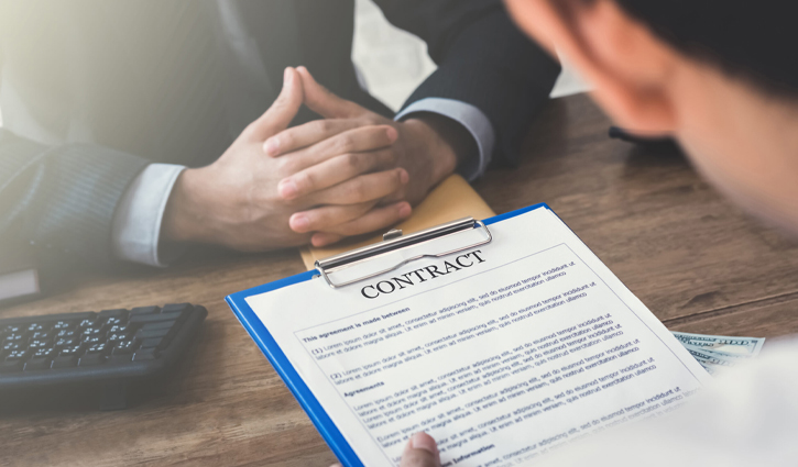 A person holding a clipboard with a contract faces another individual, their hands crossed on the desk, under the watchful eye of employers. Nearby, a calculator hints at a business negotiation where No-Poaching Agreements act as a protective shield in their discussion.