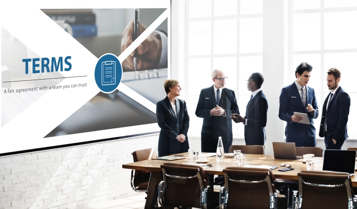 A group of business professionals stand in a modern conference room, discussing TERMS on a presentation slide. As they review Rapid Hire Solutions on the table amidst office supplies, large windows flood the space with natural light.