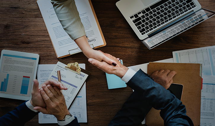 A top view of a business meeting on Mergers & Acquisitions with two people shaking hands across a table. Documents, a tablet displaying graphs, a laptop, and hands holding a pen are visible.