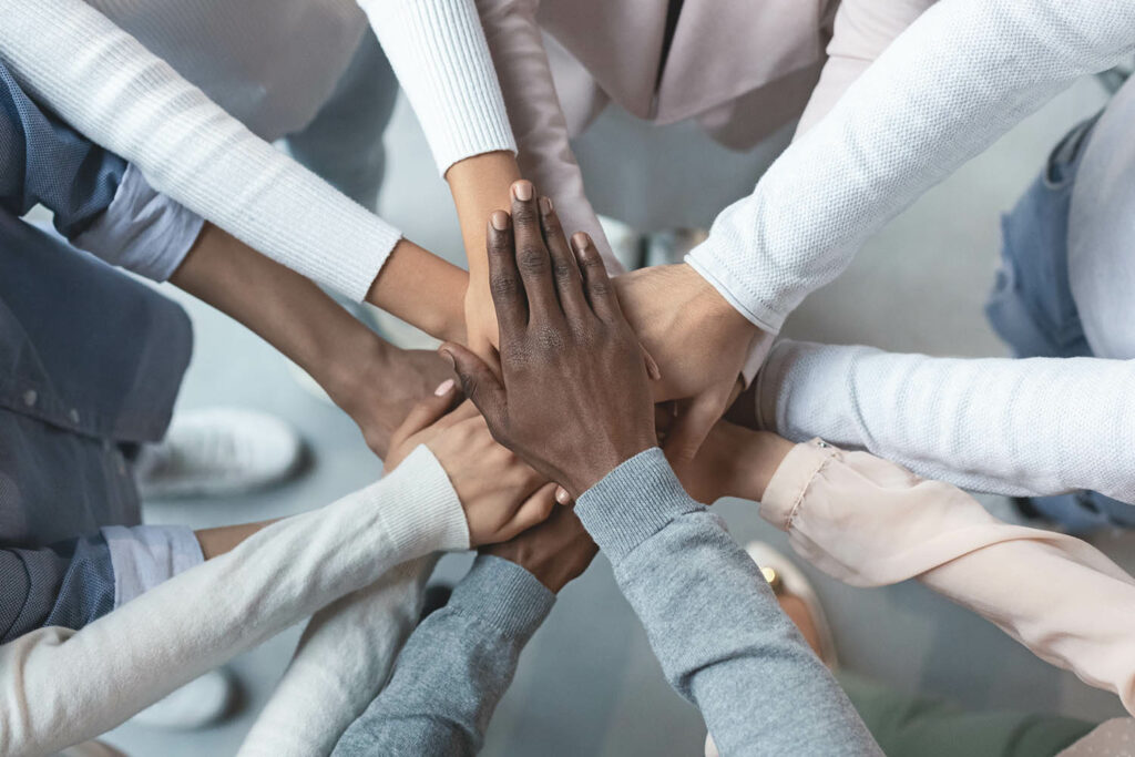 A diverse group of people stands in a circle with their hands stacked in the center, symbolizing unity and teamwork. Sporting various colored long-sleeve shirts, only their hands and arms are visible, embodying the spirit of DEI that employers strive to uphold.