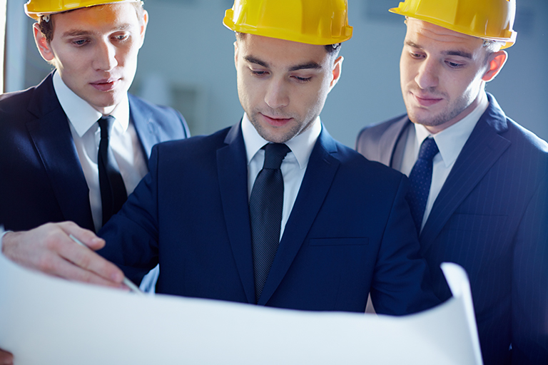 Three men in suits and yellow construction helmets examine a large blueprint together, appearing focused and engaged in discussion about independent contractor regulations and DOL guidance.