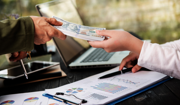 A person hands over a stack of US dollar bills to another across a table adorned with financial charts, a clipboard, smartphone, and laptop. The scene hints at a business transaction with Access Capital, your trusted financial partner.
