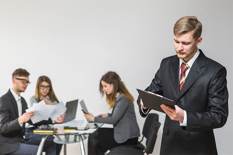 A man in a suit stands in the foreground reading a clipboard. In the background, three colleagues sit at a glass table, reviewing market documents and working on laptops in a modern office setting.