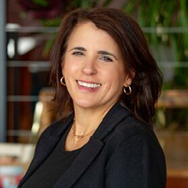 A woman with shoulder-length brown hair, wearing a black blazer and gold hoop earrings, smiles at the camera in an indoor setting with plants in the background, ready to lead a Training Series.