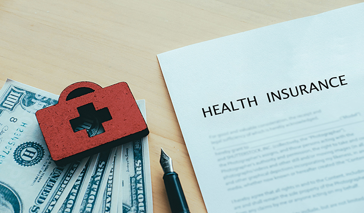 A health insurance document rests on a table beside a red medical bag icon and a stack of U.S. dollar bills, hinting at the benefits of switching to a self-funded health plan. A fountain pen lies nearby, symbolizing careful financial and insurance planning.