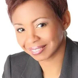 A woman with short, reddish-brown hair smiles at the camera. She is wearing a dark blazer and light makeup, ready to present a Training Series, and is posed against a plain white background.