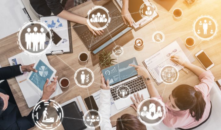 Four people gathered around a table with laptops, papers, and charts, collaborating on work. Overlaid digital icons represent teamwork and data sharing—reflecting skills-first hiring and inclusive talent pipelines in today’s staffing firms.