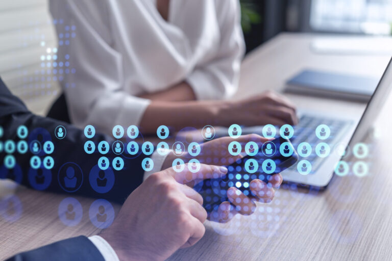 Two professionals sit at a wooden table in an office, using a smartphone and laptop. Overlaid icons of people hint at networking and communication, emphasizing growth and data-driven industry benchmarking as key elements in their drive toward profitability.