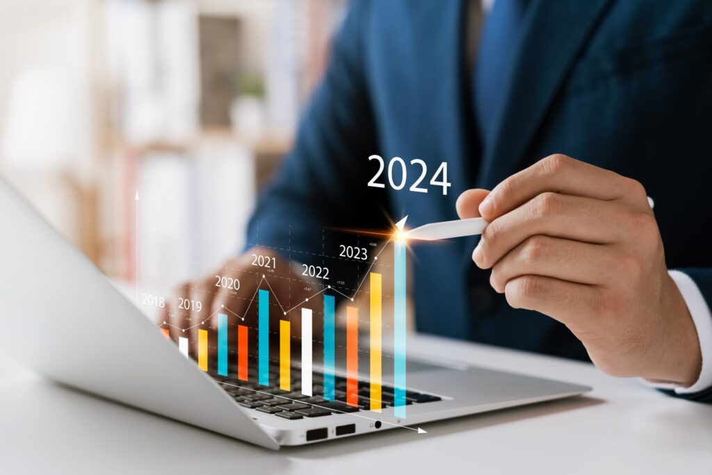 A person in a suit is using a stylus on a laptop, interacting with a digital graph. The rising bars, labeled from 2018 to 2024, highlight upward financial growth, emphasizing how the IT staffing industry with the help of advanced analytics solutions has thrived. The number 2024 is prominently featured above the bars.