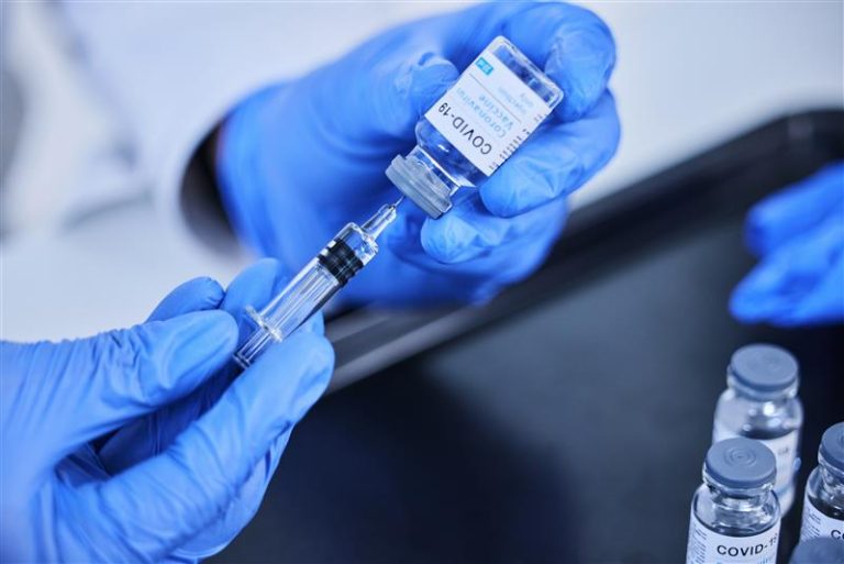 A person wearing blue gloves fills a syringe with the COVID-19 vaccine from a vial, with additional vaccine vials on an ADA-compliant black tray nearby.