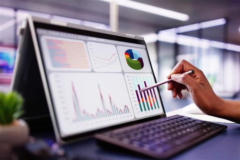 A person uses a stylus to point at colorful IT graphs and charts displayed on a tablet screen, with a keyboard in front, in a modern office setting.