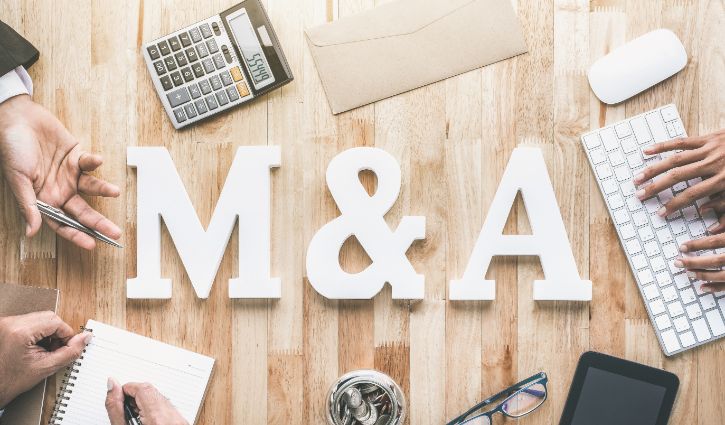 A wooden table displays large white letters M & A in the center. Around the letters, hands use a notebook, calculator, keyboard, and smartphone amidst discussions on the dynamic M&A market, suggesting a strategic business meeting setting.