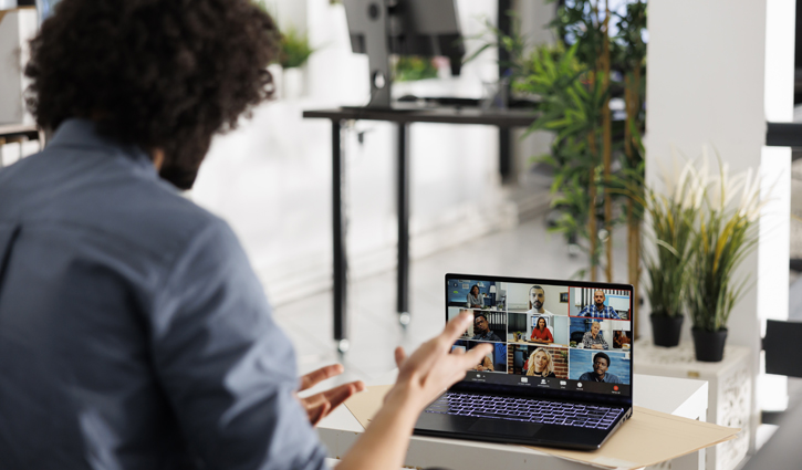 A person with curly hair participates in a video conference on a laptop, embodying the future of work. Gesturing enthusiastically while speaking, they engage with several participants onscreen. The room is modern and inviting, featuring plants, a table, and computers in the background.