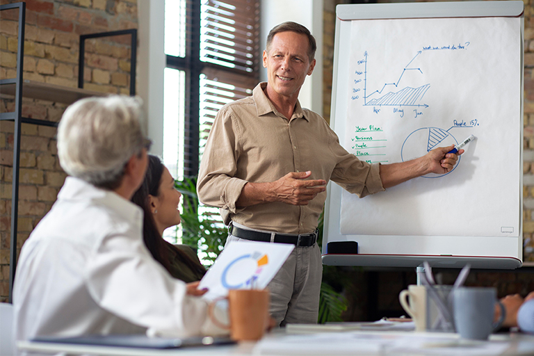 A man stands by a flip chart with market graphs and charts, presenting to three seated colleagues in a bright, modern office. He gestures toward the flip chart while the others listen and take notes.