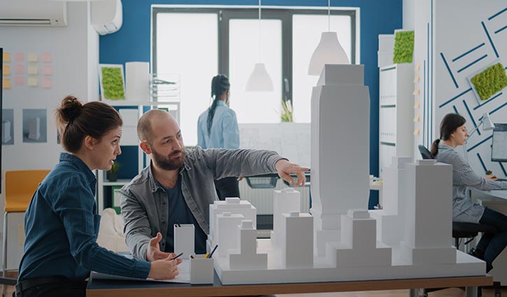 In a modern office with plants on the walls, two people discuss a scale model of a cityscape. The man points at a tall building, possibly representing the future shape of firms like UHY, while the woman listens attentively. In the background, others work at desks with computers.