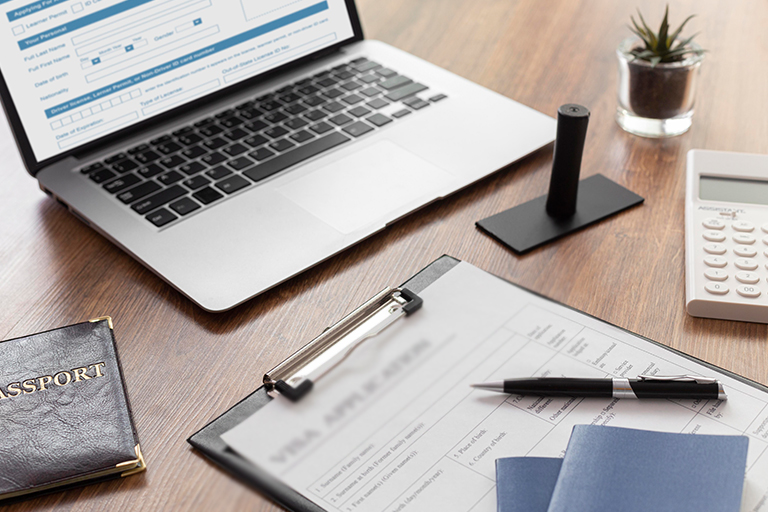 A laptop displaying a USCIS Form I-9 online sits on a wooden desk with a clipboard holding paperwork, a pen, passport, calculator, rubber stamp, and a small potted plant.