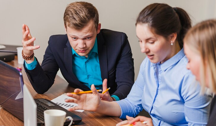 Three people are seated at a wooden table, engaged in a discussion about small business benefits. They have papers and a laptop in front of them. One person gestures with a pen, another holds papers, and a mug is also visible on the table.