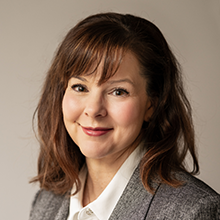 Candace Ramsahoi, with shoulder-length brown hair and bangs, wearing a grey blazer over a white collared shirt, smiles softly at the camera against a neutral background.
