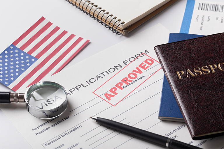 A visa application form marked approved is surrounded by a magnifying glass, a U.S. flag, a passport, airline tickets, and a notebook—highlighting the journey through student visa regulations for international student visas.