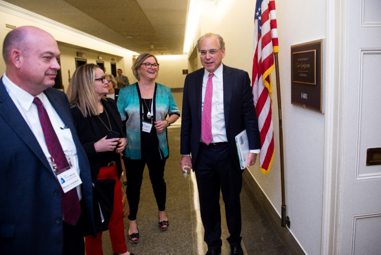 A group of four people walks through a hallway, engaged in conversation. One man holds a folder and a water bottle. An American flag is visible beside a door with a wooden sign. Theyre dressed in business attire, appearing ready to take action on their congressional campaign mission.