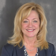 Laura Durand, a woman with blonde, shoulder-length hair, smiles at the camera. She is wearing a dark blazer over a patterned blouse, with a neutral gray background behind her.