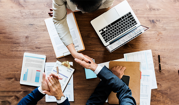 Top view of two people shaking hands over a wooden table with a laptop, clipboard, and documents. Business charts and paperwork scattered hint at an M&A meeting hitting the sweet spot. One person takes notes while observing this professional agreement between companies.