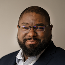 Peter Farmer, a man with short hair, a beard, and glasses, smiles at the camera. He is wearing a dark blazer and a light-colored shirt, set against a neutral background.