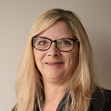 Shannon Thornbury, a woman with long blonde hair and glasses, is smiling at the camera. She is wearing a dark top and posed against a plain, light-colored background.