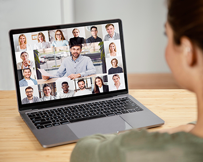 A person sits at a desk looking at a laptop screen displaying a Solution Spotlight video conference with many participants, each shown in their own small window.