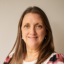 Susan Nonhof, a woman with long brown hair, smiles at the camera. She is wearing a white top with pink and black patterns and is posed against a neutral light-colored background.