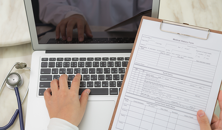 A person types on a laptop keyboard while holding a clipboard with a medical history form, mindful of the 2024 ACA reporting deadlines. A stethoscope is visible next to the laptop on the marble surface.