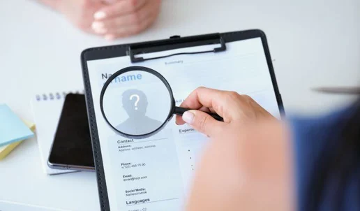 A person holds a magnifying glass over a resume on a clipboard, focusing on a section with a question mark where a profile photo would be—highlighting concerns about candidate deception. Another persons hands are visible in the background on a desk.
